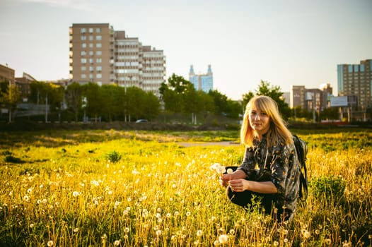 Young blonde girl in dress with shoulder bag, walking on dandelion field, against background of urban architecture, breathing relaxing in cityscape