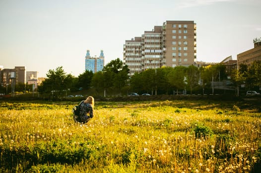Young blonde girl in dress with shoulder bag, walking on dandelion field, against background of urban architecture, breathing relaxing in cityscape