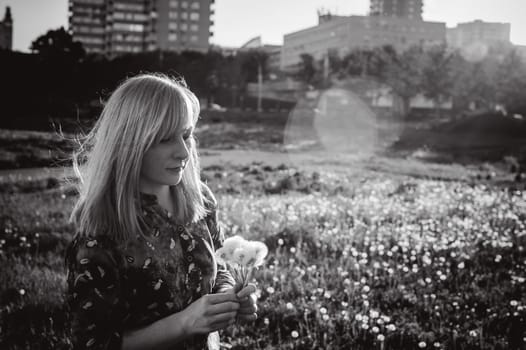 Young blonde girl in dress with shoulder bag, walking on dandelion field, against background of urban architecture, breathing relaxing in cityscape