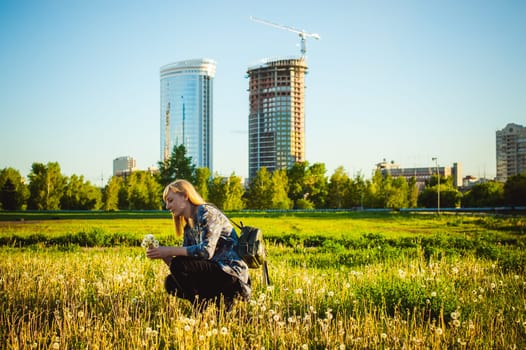 Young blonde girl in dress with shoulder bag, walking on dandelion field, against background of urban architecture, breathing relaxing in cityscape