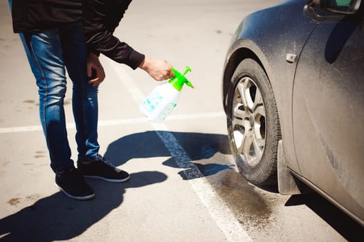 young bearded man washes his car's wheel rims, spraying water from spray, in street parking