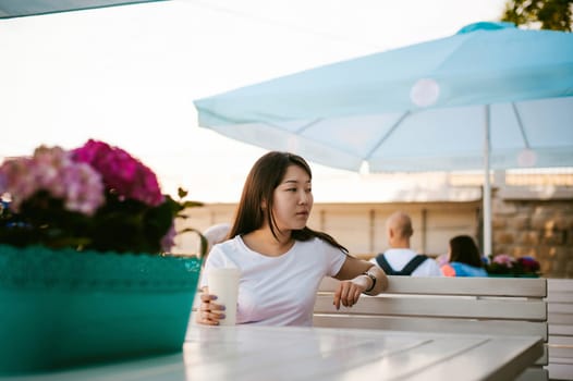 Cute asian young woman in summer cafe outdoors. girl In white T-shirt, with long hair in simple light cozy interior of restaurant Urban style, drinking coffee drink, sitting at table with flowers