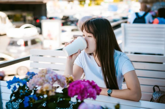 Cute asian young woman in summer cafe outdoors. girl In white T-shirt, with long hair in simple light cozy interior of restaurant Urban style, drinking coffee drink, sitting at table with flowers