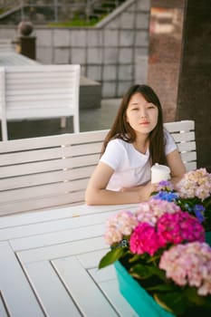Cute asian young woman in summer cafe outdoors. girl In white T-shirt, with long hair in simple light cozy interior of restaurant Urban style, drinking coffee drink, sitting at table with flowers