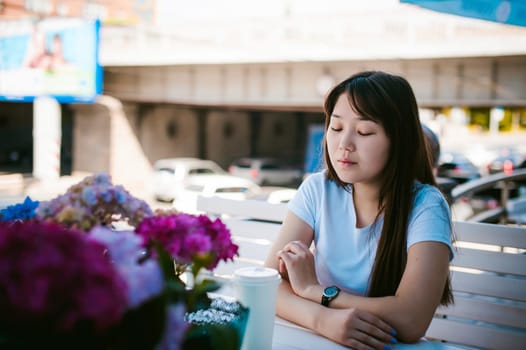 Cute asian young woman in summer cafe outdoors. girl In white T-shirt, with long hair in simple light cozy interior of restaurant Urban style, drinking coffee drink, sitting at table with flowers