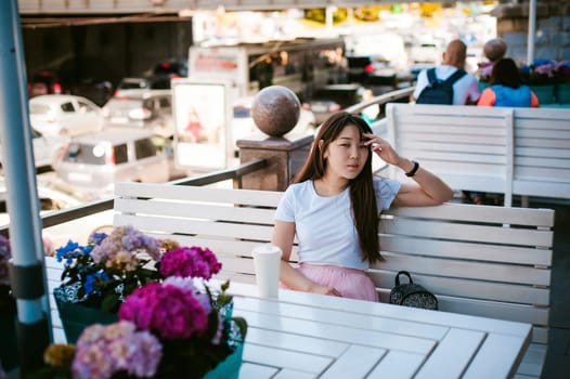 Cute asian young woman in summer cafe outdoors. girl In white T-shirt, with long hair in simple light cozy interior of restaurant Urban style, drinking coffee drink, sitting at table with flowers