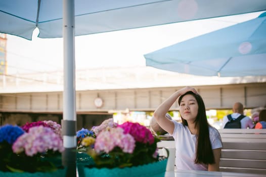 Cute asian young woman in summer cafe outdoors. girl In white T-shirt, with long hair in simple light cozy interior of restaurant Urban style, drinking coffee drink, sitting at table with flowers