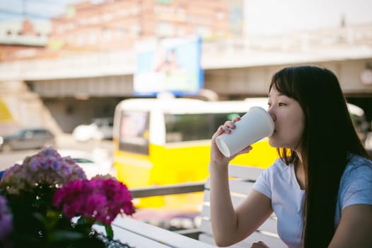 Cute asian young woman in summer cafe outdoors. girl In white T-shirt, with long hair in simple light cozy interior of restaurant Urban style, drinking coffee drink, sitting at table with flowers