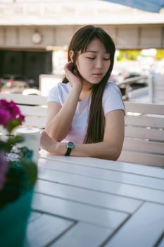 Cute asian young woman in summer cafe outdoors. girl In white T-shirt, with long hair in simple light cozy interior of restaurant Urban style, drinking coffee drink, sitting at table with flowers