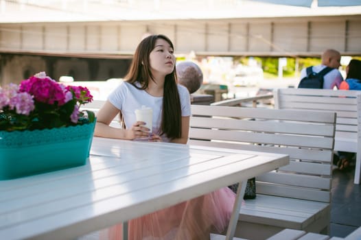 Cute asian young woman in summer cafe outdoors. girl In white T-shirt, with long hair in simple light cozy interior of restaurant Urban style, drinking coffee drink, sitting at table with flowers