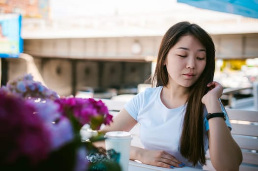Cute asian young woman in summer cafe outdoors. girl In white T-shirt, with long hair in simple light cozy interior of restaurant Urban style, drinking coffee drink, sitting at table with flowers