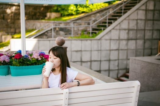Cute asian young woman in summer cafe outdoors. girl In white T-shirt, with long hair in simple light cozy interior of restaurant Urban style, drinking coffee drink, sitting at table with flowers