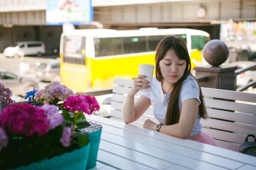 Cute asian young woman in summer cafe outdoors. girl In white T-shirt, with long hair in simple light cozy interior of restaurant Urban style, drinking coffee drink, sitting at table with flowers