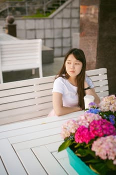 Cute asian young woman in summer cafe outdoors. girl In white T-shirt, with long hair in simple light cozy interior of restaurant Urban style, drinking coffee drink, sitting at table with flowers