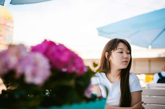 Cute asian young woman in summer cafe outdoors. girl In white T-shirt, with long hair in simple light cozy interior of restaurant Urban style, drinking coffee drink, sitting at table with flowers