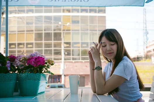 Cute asian young woman in summer cafe outdoors. girl In white T-shirt, with long hair in simple light cozy interior of restaurant Urban style, drinking coffee drink, sitting at table with flowers