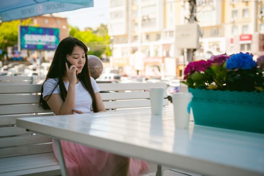 Cute asian young woman in summer cafe outdoors. girl In white T-shirt, with long hair in simple light cozy interior of restaurant Urban style, Communicates on a cell phone holding it in his hands