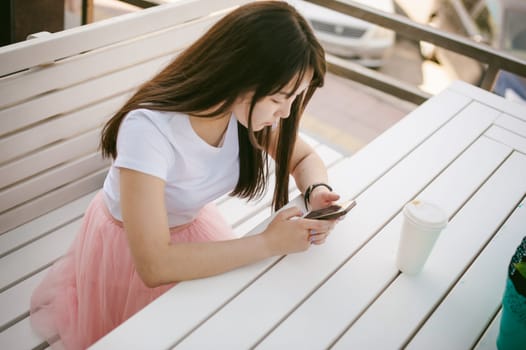 Cute asian young woman in summer cafe outdoors. girl In white T-shirt, with long hair in simple light cozy interior of restaurant Urban style, Communicates on a cell phone holding it in his hands