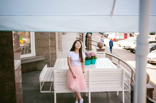 Cute asian young woman in summer cafe outdoors. girl In white T-shirt, with long hair in simple light cozy interior of restaurant Urban style