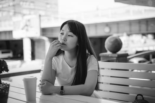 Cute asian young woman in summer cafe outdoors. girl In white T-shirt, with long hair in simple light cozy interior of restaurant Urban style, drinking coffee drink, sitting at table with flowers