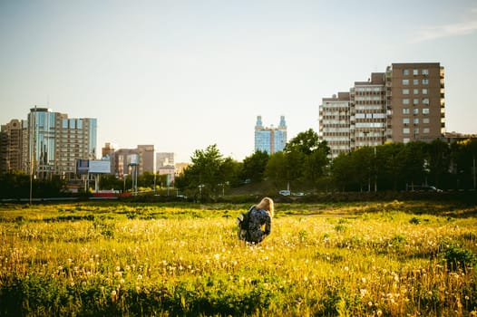 Young blonde girl in dress with shoulder bag, walking on dandelion field, against background of urban architecture, breathing relaxing in cityscape