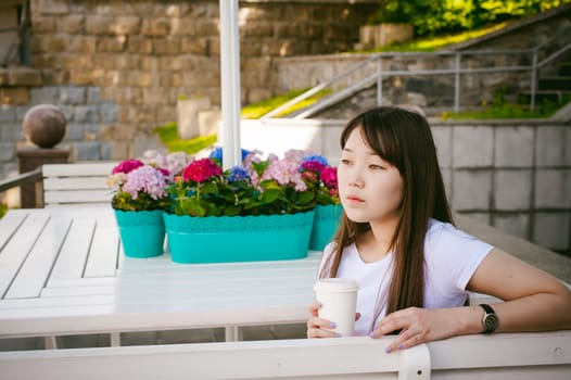 Cute asian young woman in summer cafe outdoors. girl In white T-shirt, with long hair in simple light cozy interior of restaurant Urban style, drinking coffee drink, sitting at table with flowers