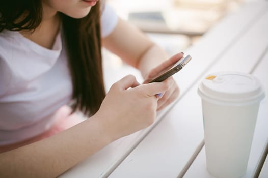 Cute asian young woman in summer cafe outdoors. girl In white T-shirt, with long hair in simple light cozy interior of restaurant Urban style, Communicates on a cell phone holding it in his hands