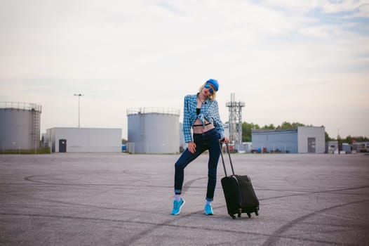 young slim woman dressed in blue checked shirt Sunglasses, cap and jeans, Will depart at dawn on summer day on freedom trip with black travel suitcase