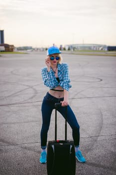 young slim woman dressed in blue checked shirt Sunglasses, cap and jeans, Will depart at dawn on summer day on freedom trip with black travel suitcase