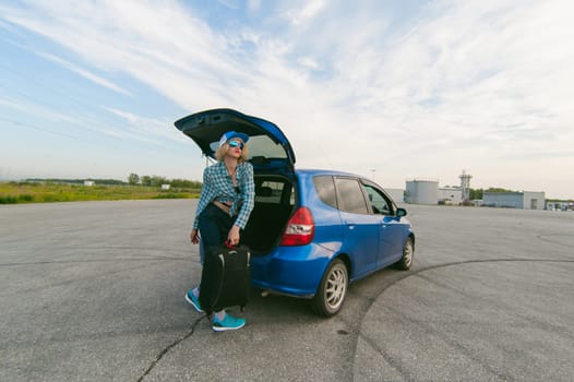 young slim woman dressed in blue checked shirt, cap and jeans, Will depart at dawn on summer day on freedom trip with black travel suitcase. Takes out a bag from the car