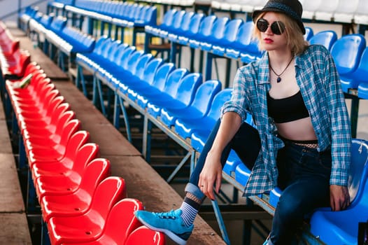 young slim woman dressed in blue checked shirt, cap and jeans, Will depart at dawn on summer day Sits spectator places of the fans Smokes a cigarette