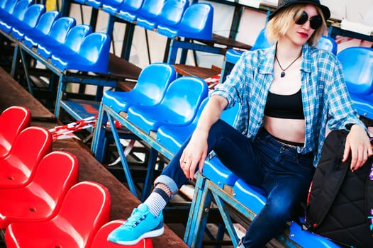 young slim woman dressed in blue checked shirt, cap and jeans, Will depart at dawn on summer day Sits spectator places of the fans Smokes a cigarette