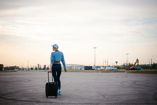young slim woman dressed in blue checked shirt, cap and jeans, Will depart at dawn on summer day on freedom trip with black travel suitcase