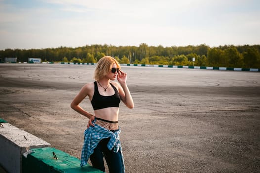 young slim woman dressed in blue checked shirt Sunglasses, cap and jeans, Pantyhose in the net and topic. Will depart at dawn on summer day. Sits on concrete block, bump of race track