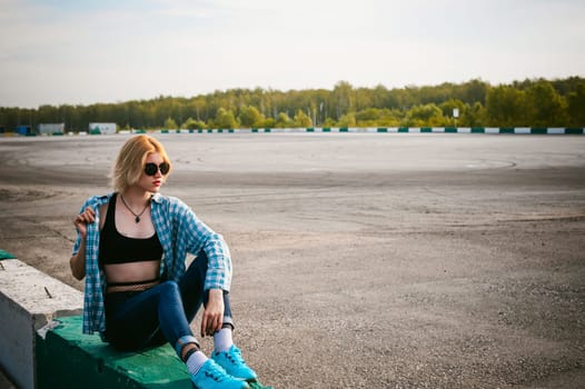 young slim woman dressed in blue checked shirt Sunglasses, cap and jeans, Pantyhose in the net and topic. Will depart at dawn on summer day. Sits on concrete block, bump of race track