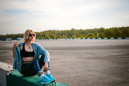 young slim woman dressed in blue checked shirt Sunglasses, cap and jeans, Pantyhose in the net and topic. Will depart at dawn on summer day. Sits on concrete block, bump of race track