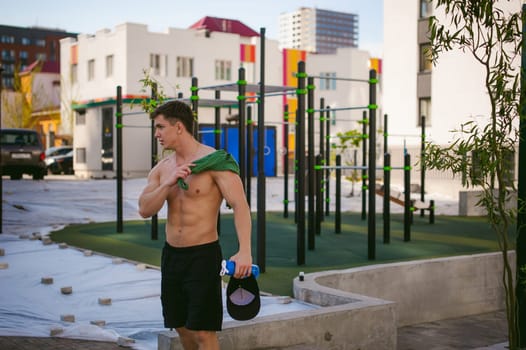 Handsome sexy male bodybuilder athlete man with naked torso, Poses showing a powerful muscular body standing near a green grid fence sports field in courtyard. Healthy lifestyle concept.