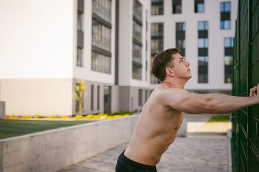 Handsome sexy male bodybuilder athlete man with naked torso, Poses showing a powerful muscular body standing near a green grid fence sports field in courtyard. Healthy lifestyle concept.