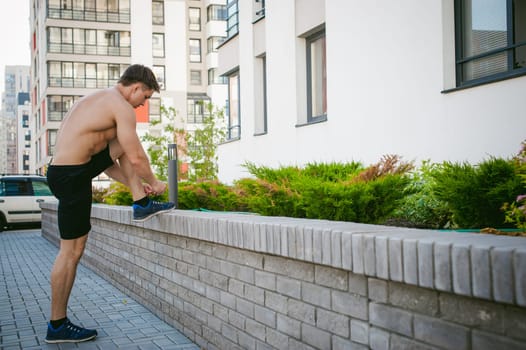 Handsome sexy male bodybuilder athlete man with naked torso, running in morning, outdoors. ties up shoelaces on sneakers. Healthy lifestyle concept.