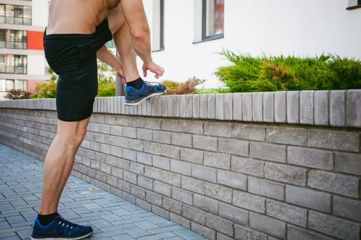 Handsome sexy male bodybuilder athlete man with naked torso, running in morning, outdoors. ties up shoelaces on sneakers. Healthy lifestyle concept.