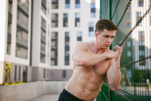 Handsome sexy male bodybuilder athlete man with naked torso, Poses showing a powerful muscular body standing near a green grid fence sports field in courtyard. Healthy lifestyle concept.