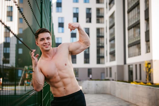 Handsome sexy male bodybuilder athlete man with naked torso, Poses showing a powerful muscular body standing near a green grid fence sports field in courtyard. Healthy lifestyle concept.