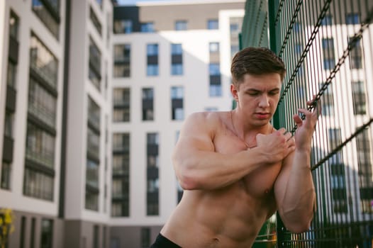 Handsome sexy male bodybuilder athlete man with naked torso, Poses showing a powerful muscular body standing near a green grid fence sports field in courtyard. Healthy lifestyle concept.