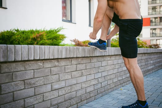 Handsome sexy male bodybuilder athlete man with naked torso, running in morning, outdoors. ties up shoelaces on sneakers. Healthy lifestyle concept.