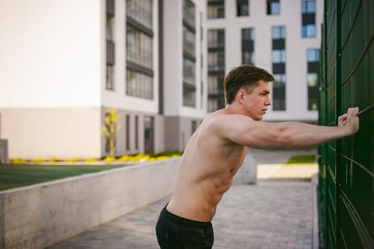 Handsome sexy male bodybuilder athlete man with naked torso, Poses showing a powerful muscular body standing near a green grid fence sports field in courtyard. Healthy lifestyle concept.