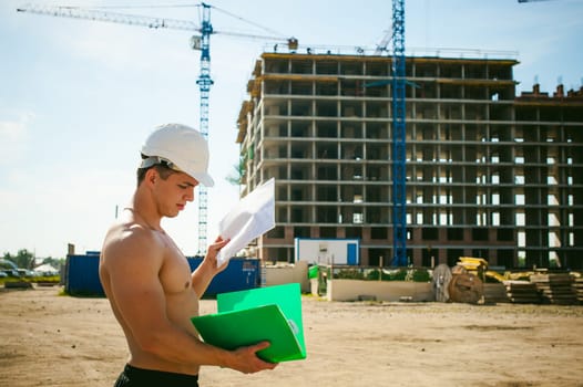 Young sexy male bodybuilder athlete with a bare torso. Manager engineer builder in white protective helmet stands on background of construction of building and construction cranes with documentation