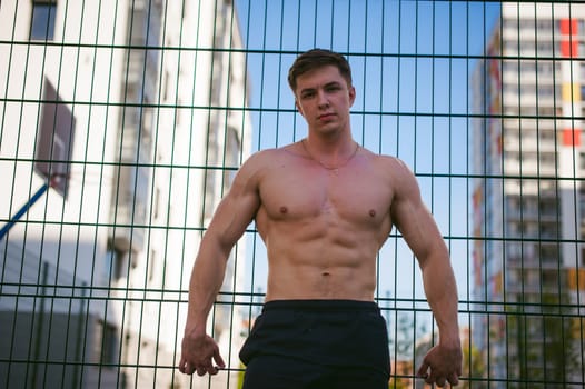 Handsome sexy male bodybuilder athlete man with naked torso, Poses showing a powerful muscular body standing near a green grid fence sports field in courtyard. Healthy lifestyle concept.