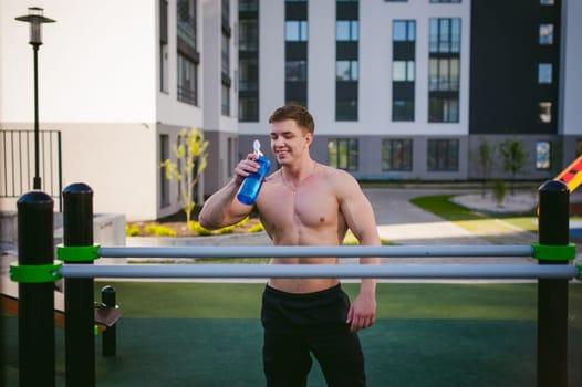 Handsome sexy male bodybuilder athlete man doing crossfit workout in athletic facilities on sunny morning outdoors. Healthy lifestyle concept. Rest after exercise, drinking water from a bottle