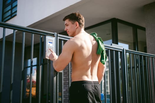 Handsome sexy male athlete man with naked torso, stands near gate, calling intercom to open door, after morning run in open air in urban courtyards, an early sunny summer morning