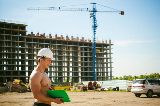 Young sexy male bodybuilder athlete with a bare torso. Manager engineer builder in white protective helmet stands on background of construction of building and construction cranes with documentation